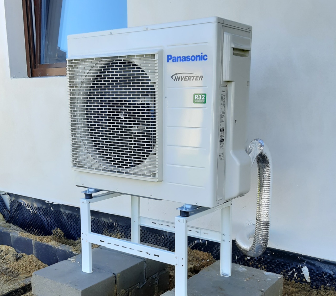 Zewnętrzna jednostka pompy ciepła Panasonic Inverter R32 na białym stelażu, zamontowana przy jasnej ścianie budynku z widocznym oknem i folią kubełkową przy fundamencie.