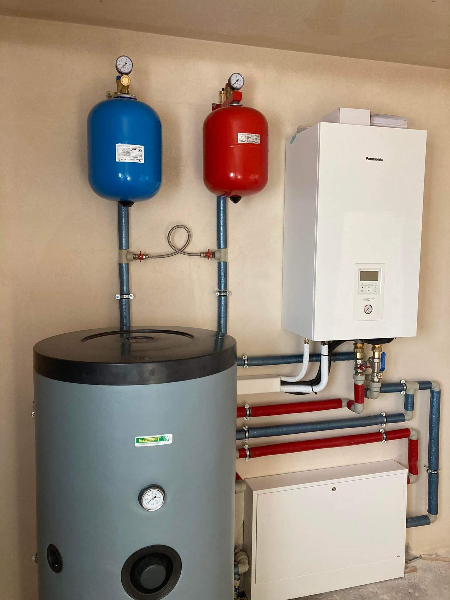 Instalacja grzewcza z zbiornikiem akumulacyjnym, dwoma naczyniami wzbiorczymi (niebieskim i czerwonym) z manometrami oraz wiszącym kotłem gazowym Panasonic, widoczne rury w izolacji termicznej.