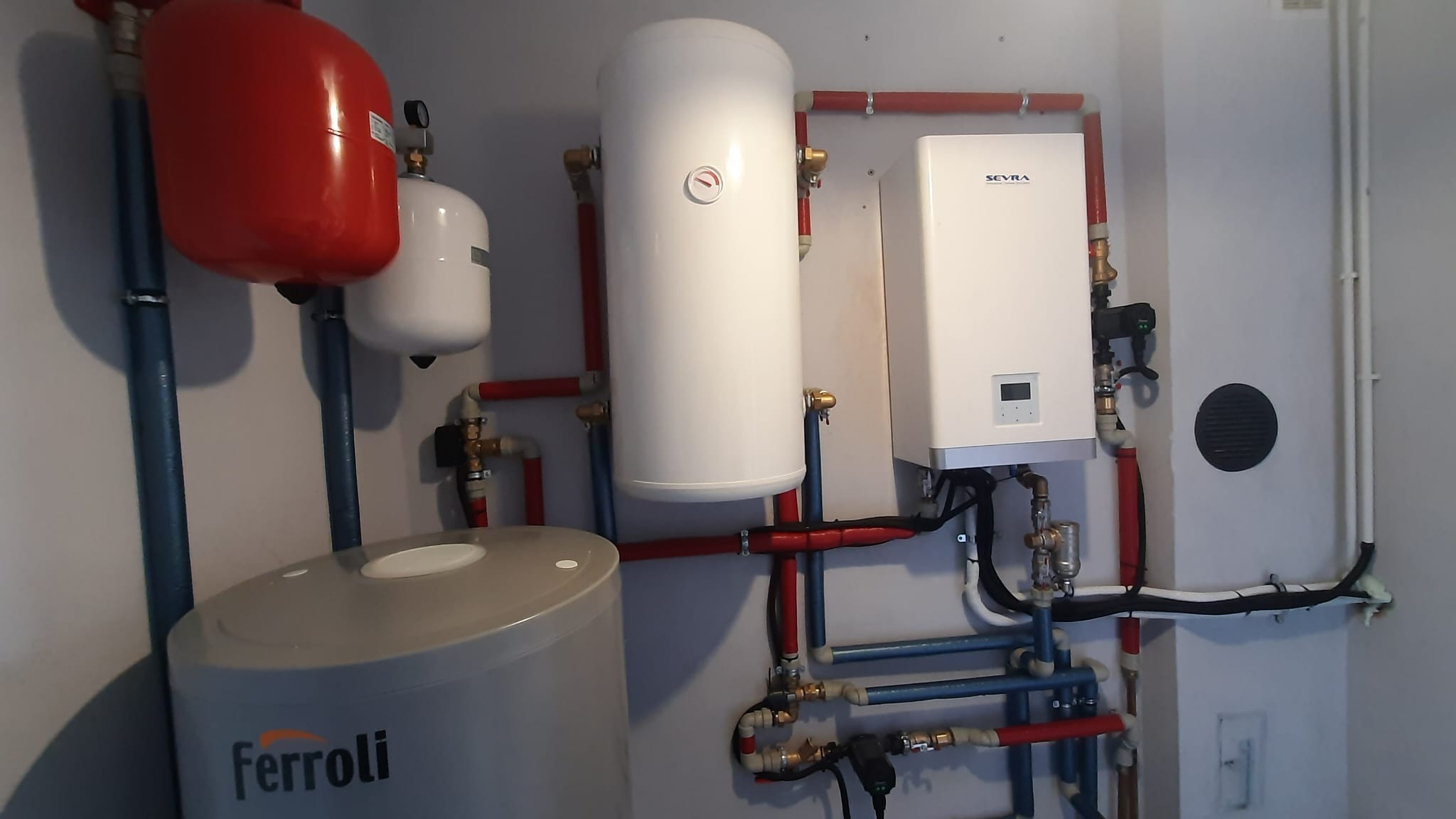 Instalacja grzewcza w kotłowni z zbiornikami Ferroli i urządzeniem Sevra, widoczne rury w kolorach czerwonym i niebieskim.