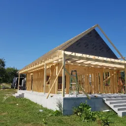 Konstrukcja szkieletowa domu w trakcie budowy, widoczne drewniane ramy ścian i dachu z częściowo zamontowaną membraną, betonowa podstawa z schodami, pracownik na drabinie w tle.