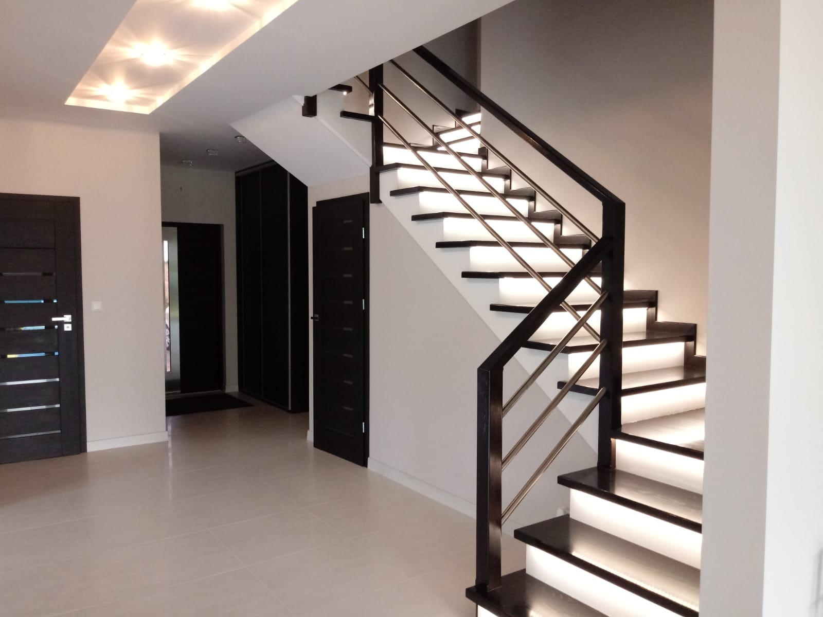 Nowoczesne schody drewniane z podświetleniem LED w Białymstoku, z ciemnymi stopniami i metalową balustradą, widoczne drzwi w kolorze wenge.