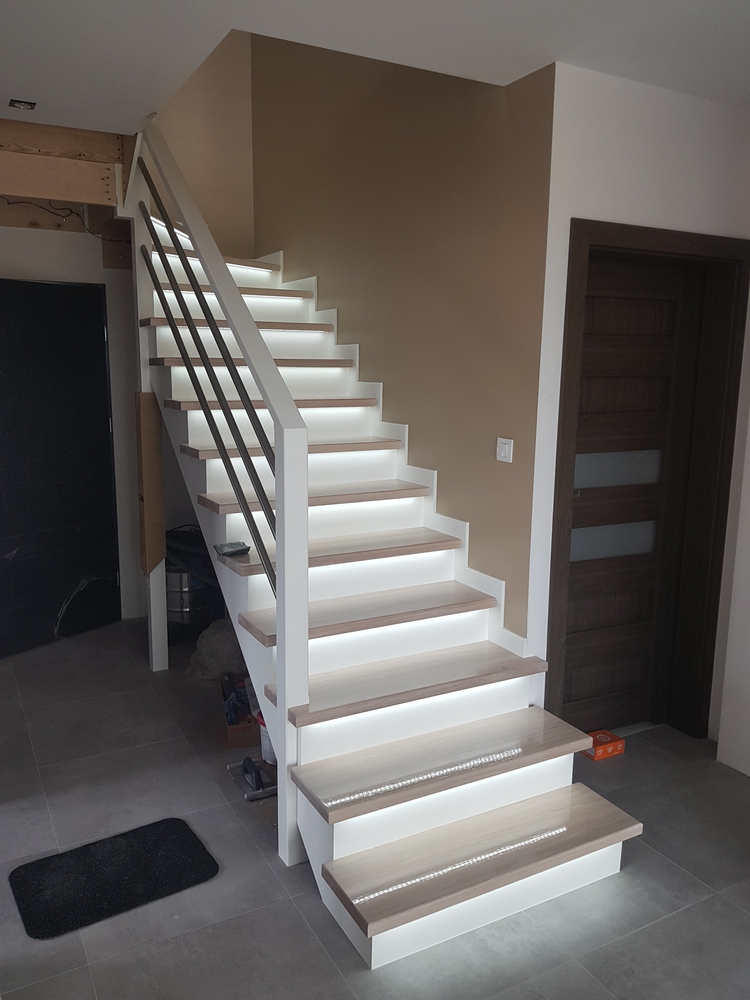 Nowoczesne schody z podświetleniem LED wzdłuż stopni, metalowe balustrady, białe boki stopni, widoczna cześć instalacji elektrycznej na suficie, szara podłoga.