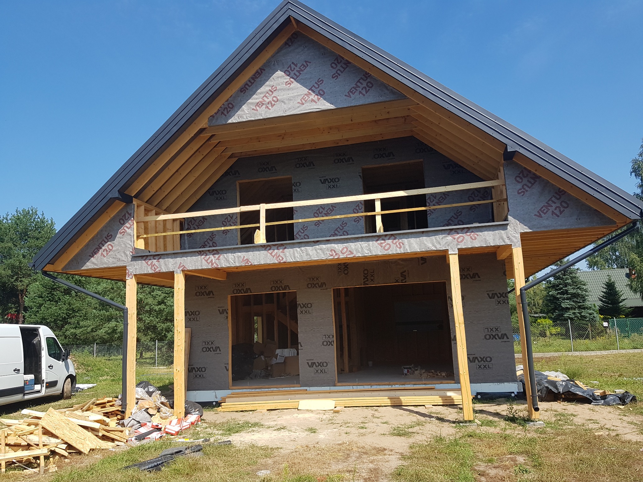 Wznoszenie konstrukcji domu z drewnianym szkieletem, widoczne drewniane elementy konstrukcyjne, membrana dachowa z nadrukiem, częściowo zamontowane orynnowanie, na pierwszym planie porozrzucane...