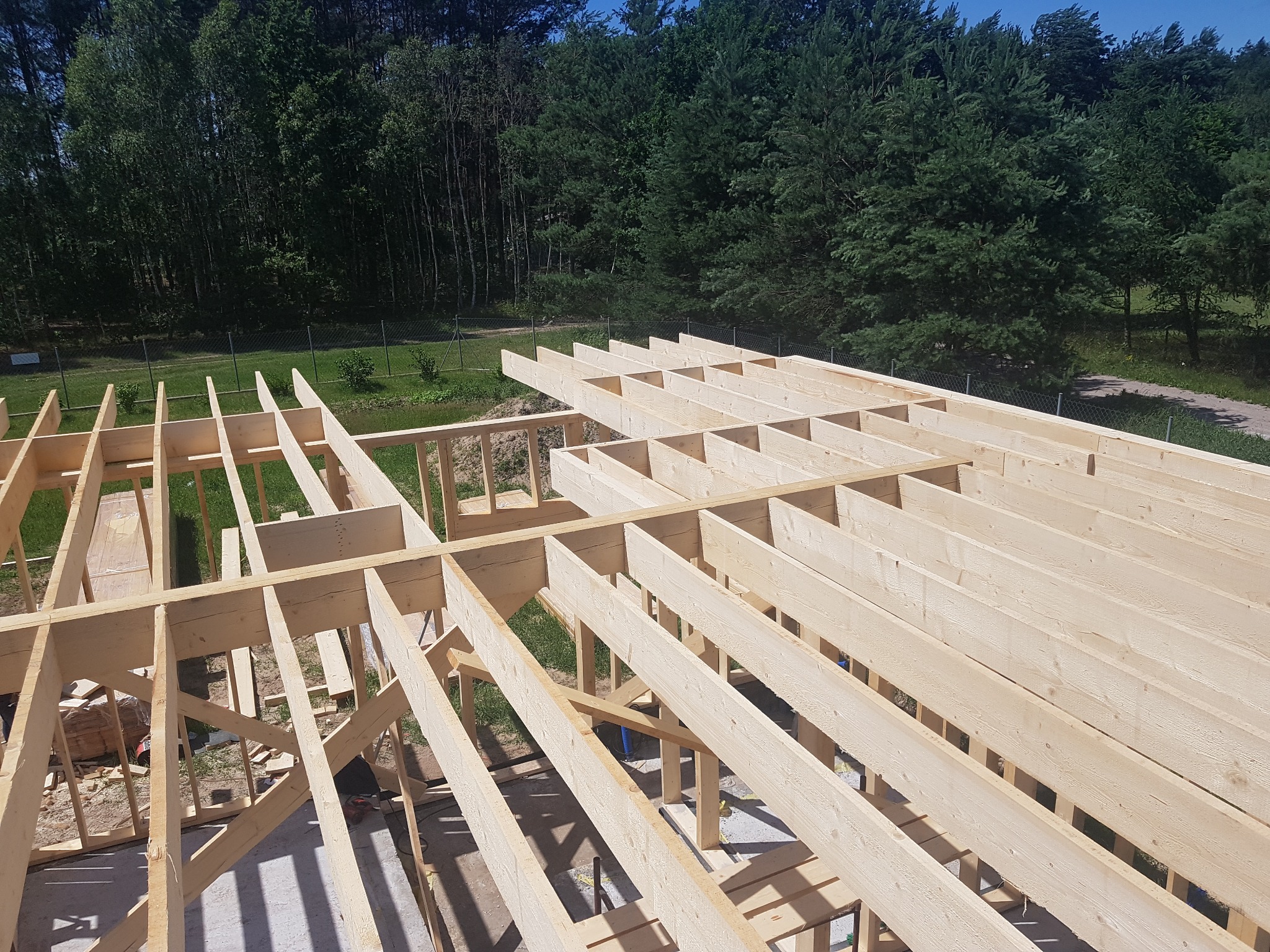 Konstrukcja drewniana domu w trakcie budowy, widok z góry na szkielet dachu i ścian, otoczona zielonym lasem w tle.