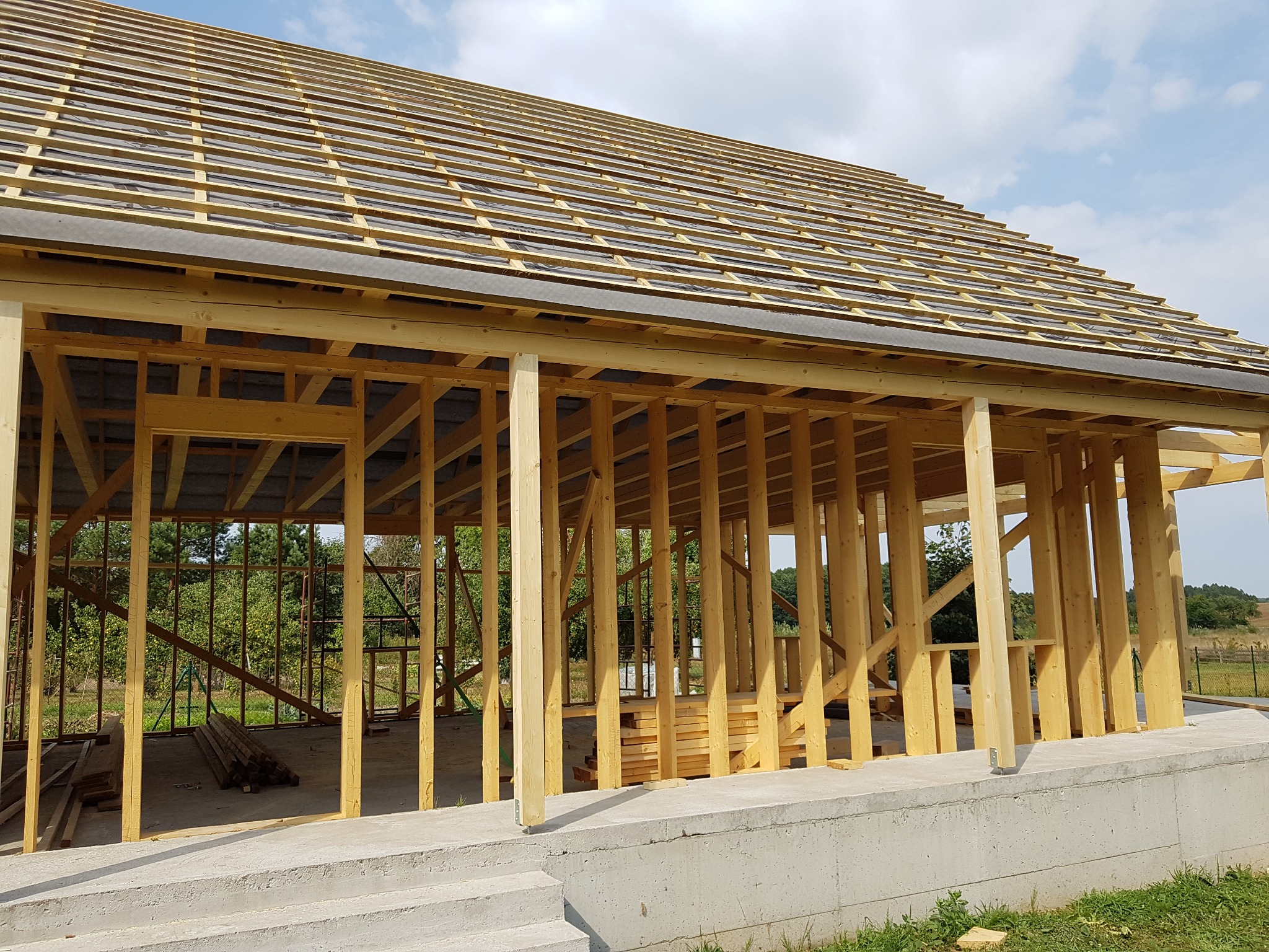 Konstrukcja domu w trakcie budowy: drewniany szkielet z widocznym deskowaniem dachu i betonową podstawą, otoczona zielenią.