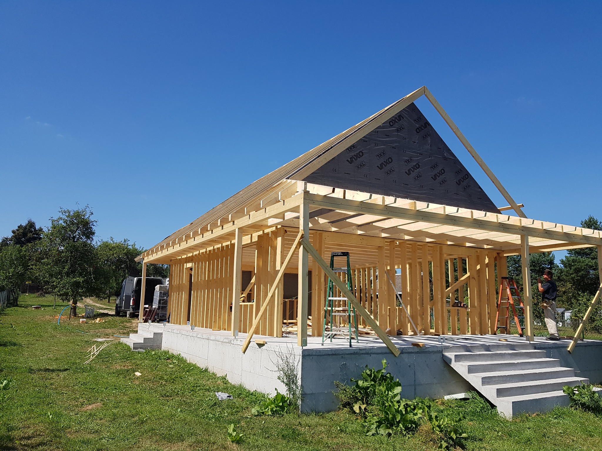 Konstrukcja szkieletowa domu w trakcie budowy, widoczne drewniane ramy ścian i dachu z częściowo zamontowaną membraną, betonowa podstawa z schodami, pracownik na drabinie w tle.