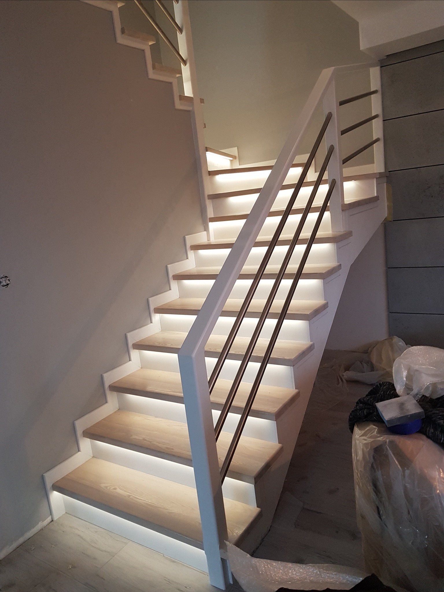 Nowoczesne schody drewniane z podświetleniem LED na każdym stopniu i metalową balustradą z białymi wspornikami, widoczne ślady prac wykończeniowych.