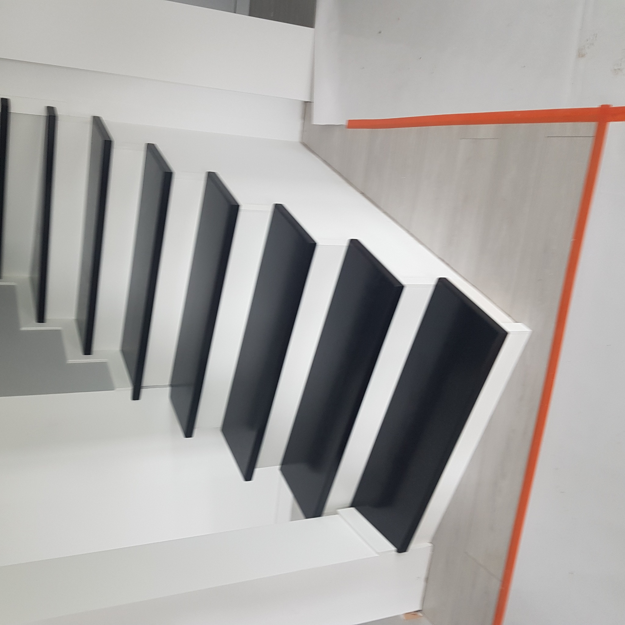 Nowoczesne schody z białymi stopniami i czarnymi podstopnicami, widok z góry, częściowo widoczna podłoga zabezpieczona pomarańczową taśmą malarską.