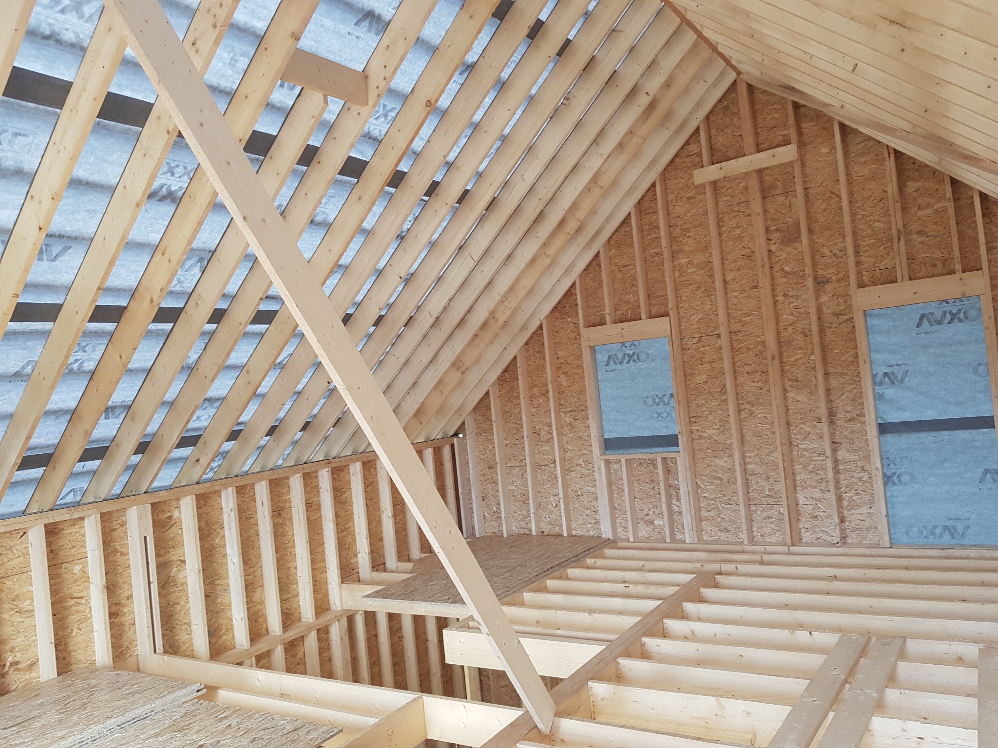 Wnętrze w trakcie budowy, widoczne drewniane elementy konstrukcyjne ścian, podłogi i dachu, z fragmentarycznie zamontowaną płytą OSB i folią paroizolacyjną na zewnątrz.