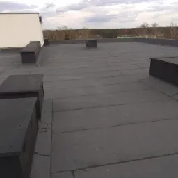 Płaski dach pokryty papą termozgrzewalną z czarnymi elementami wentylacyjnymi i kominkami, widok z góry, w tle drzewa i pochmurne niebo.