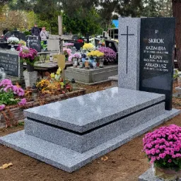 Granitowy nagrobek w odcieniach szarości z czarną tablicą, ozdobiony kwiatami w doniczkach na cmentarzu, w tle inne groby i drzewa.