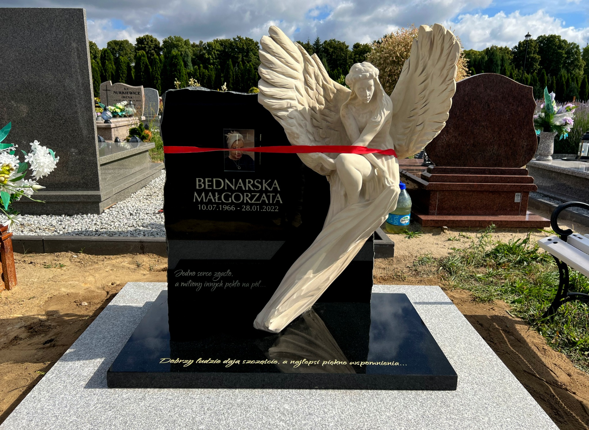 Czarny nagrobek z jasną rzeźbą anioła, przewiązany czerwoną wstążką, z wygrawerowanym imieniem i nazwiskiem oraz datami urodzenia i śmierci, umieszczony na cmentarzu.