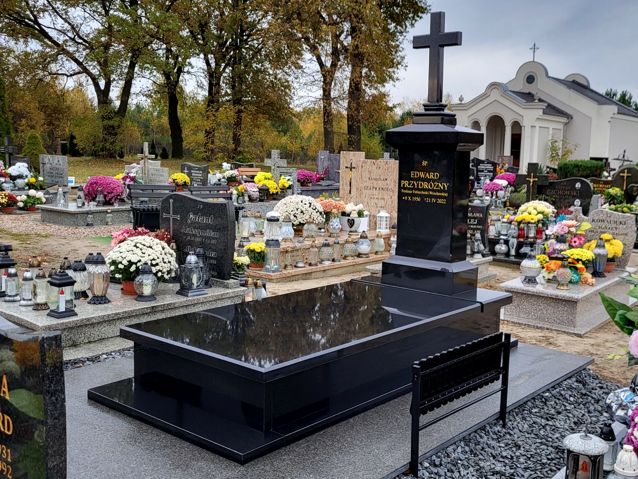 Czarny, błyszczący nagrobek z krzyżem na cmentarzu, z wygrawerowanym imieniem i nazwiskiem oraz datami urodzenia i śmierci, otoczony innymi nagrobkami, kwiatami i zniczami.