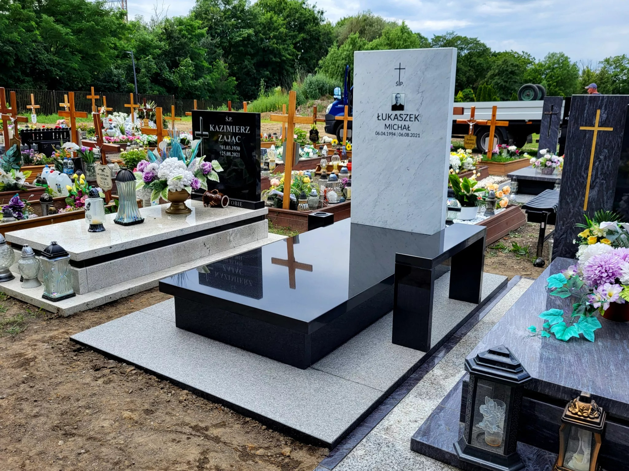 Nowoczesny, czarny nagrobek z elementami białego marmuru i siedziskiem na cmentarzu, z widocznym odbiciem krzyża na polerowanej powierzchni.