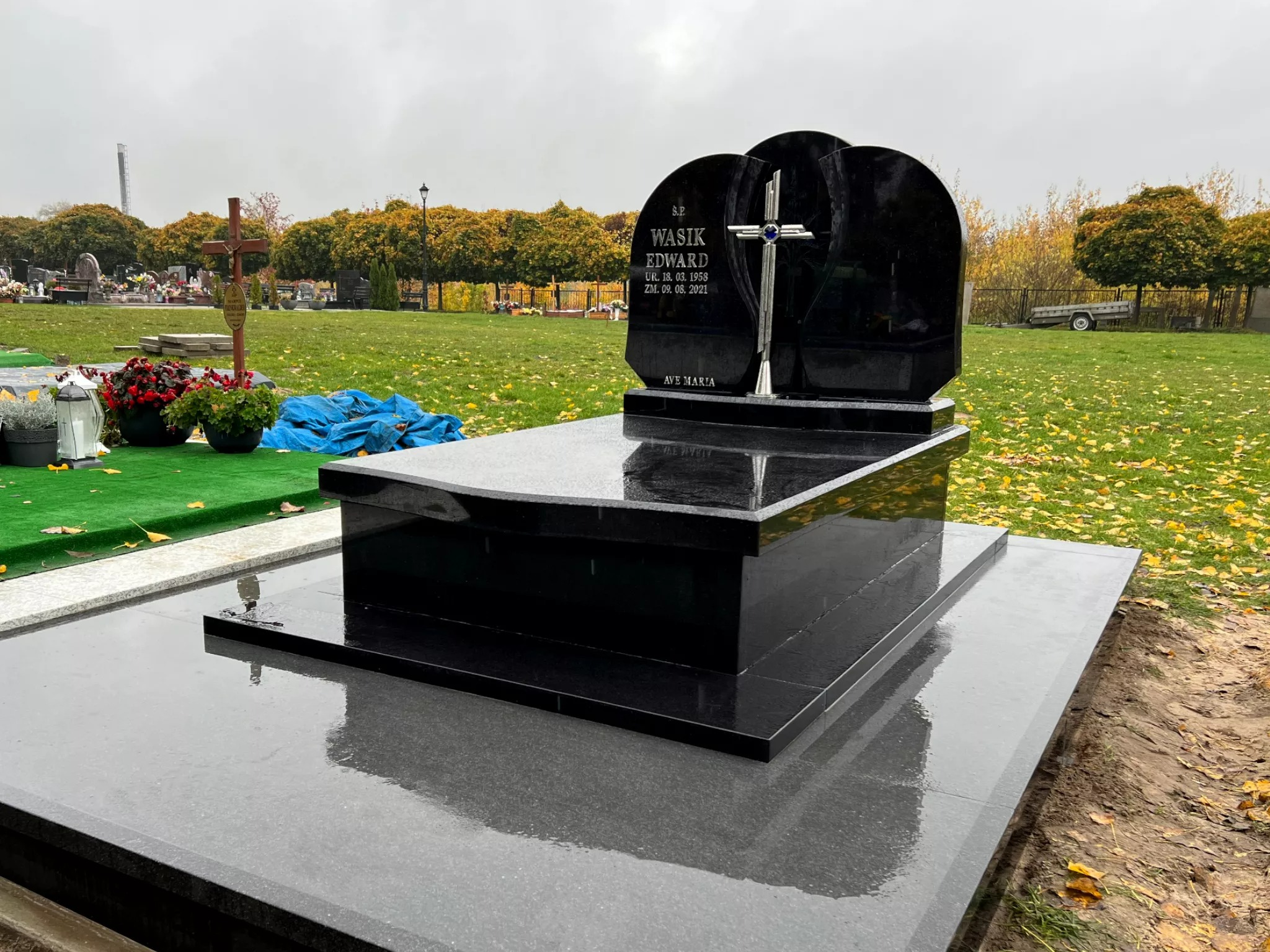 Czarny, błyszczący nagrobek z wygrawerowanym imieniem i nazwiskiem Wasik Edward oraz datami urodzenia i śmierci, z metalowym krzyżem, na cmentarzu z zielonym trawnikiem i drzewami w tle.