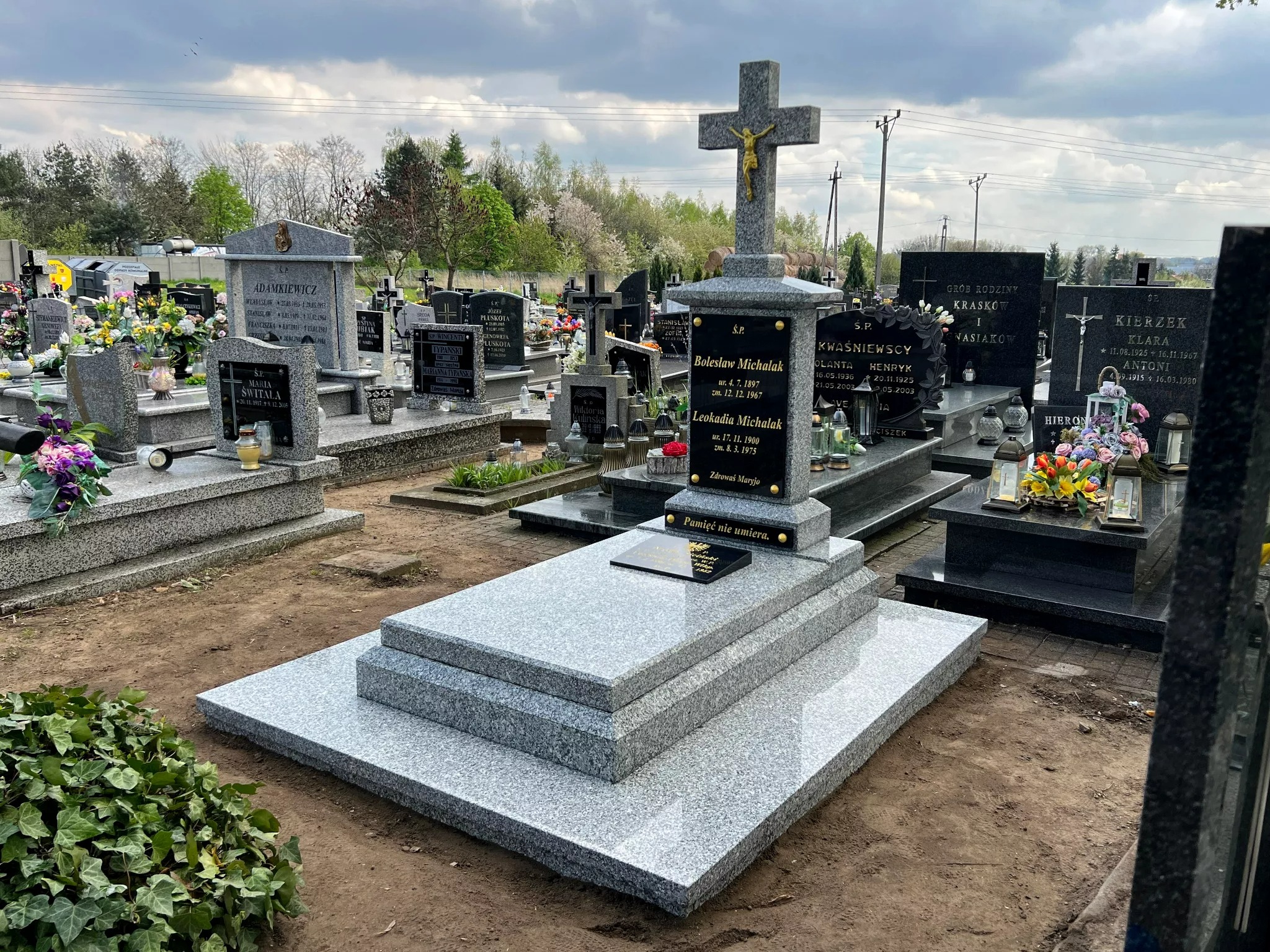 Nowy, jasnoszary, granitowy nagrobek z krzyżem na cmentarzu, z inskrypcjami i tabliczką 'Pamięć nie umiera', otoczony innymi nagrobkami i zielenią.