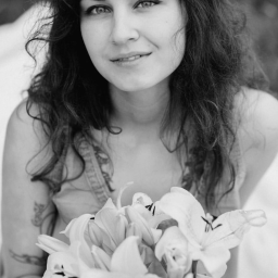 Anastazja Belska - Portret młodej kobiety z ciemnymi, kręconymi włosami, trzymającej bukiet lilii w czarno-białej tonacji, z widocznym tatuażem na ramieniu i delikatnym uśmiechem.