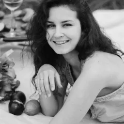 Czarno-białe zdjęcie uśmiechniętej kobiety z kręconymi włosami leżącej na kocu piknikowym z owocami i kieliszkiem wina w tle.