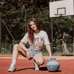 Anastazja Belska - Młoda kobieta w sportowym stroju kuca na czerwonym boisku do koszykówki, opierając dłoń na niebieskiej piłce Spalding NBA, w tle widoczny kosz i zielone drzewa.