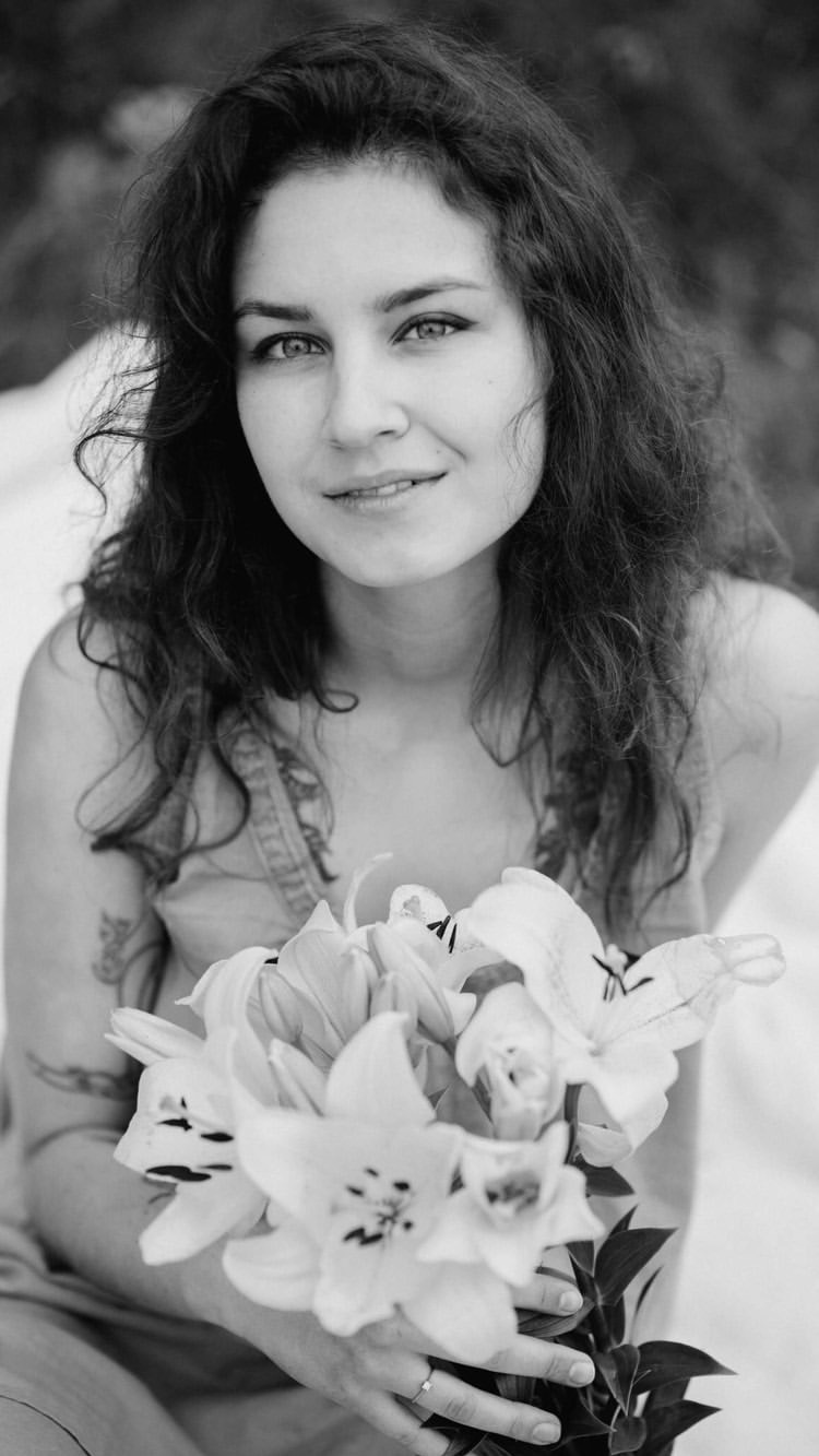 Portret młodej kobiety z ciemnymi, kręconymi włosami, trzymającej bukiet lilii w czarno-białej tonacji, z widocznym tatuażem na ramieniu i delikatnym uśmiechem.