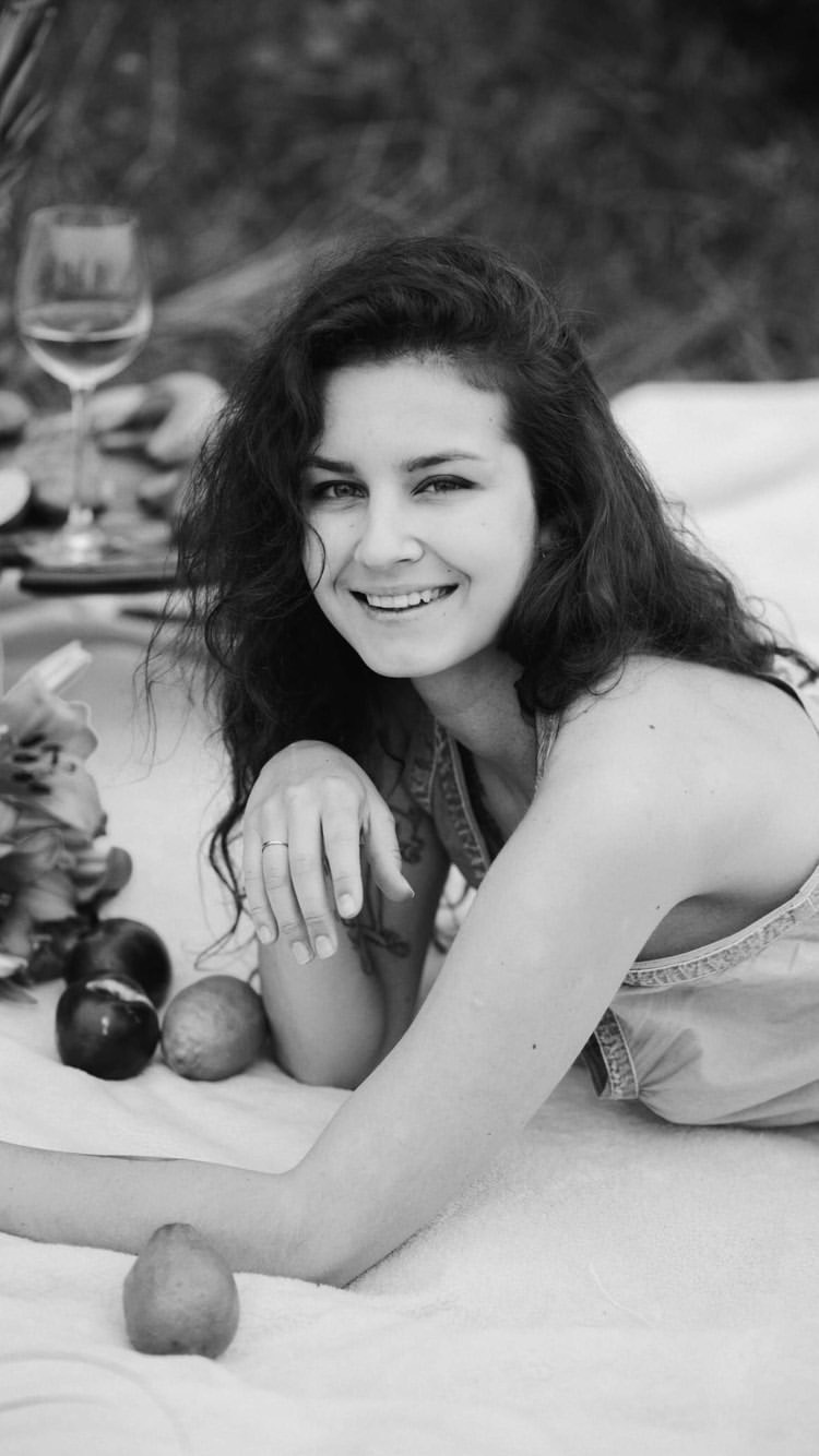 Czarno-białe zdjęcie uśmiechniętej kobiety z kręconymi włosami leżącej na kocu piknikowym z owocami i kieliszkiem wina w tle.