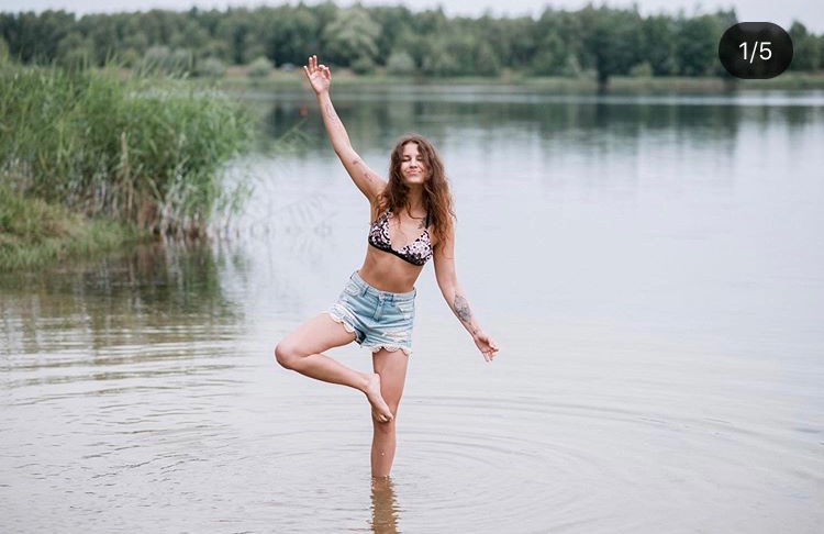 Kobieta w bikini i dżinsowych szortach stoi w jeziorze na jednej nodze, z podniesioną ręką, w tle woda i zieleń.