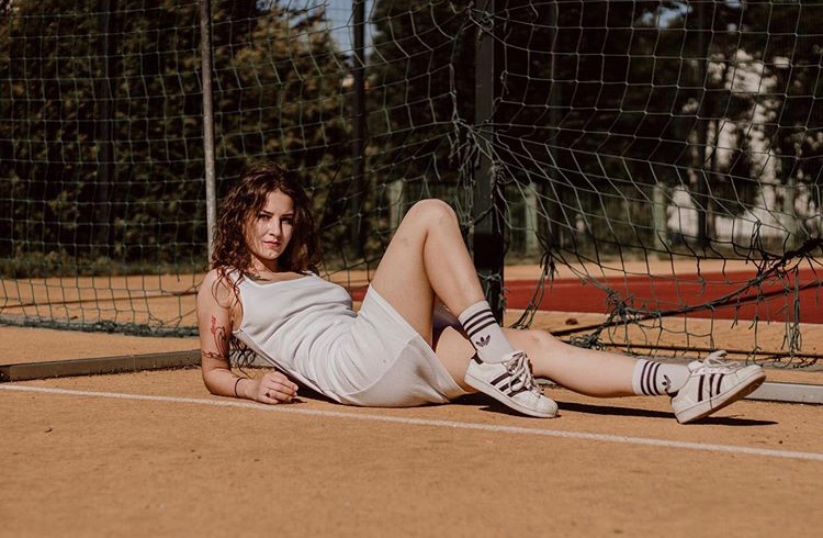 Portret młodej kobiety w białej sukience i sportowych butach, leżącej na pomarańczowym boisku do tenisa, z siatką w tle, w naturalnym świetle.