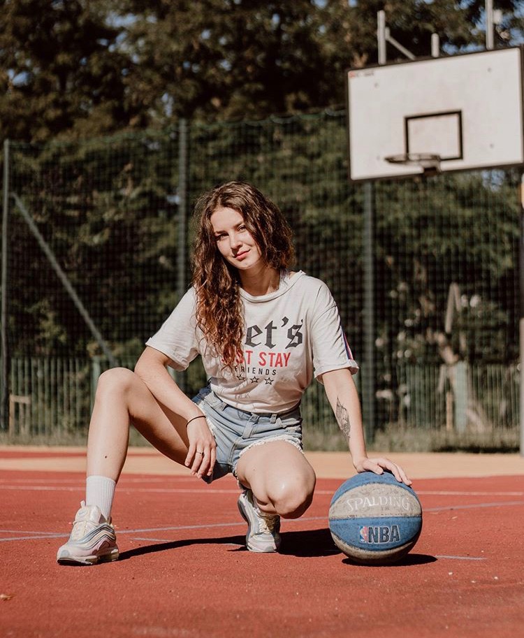 Młoda kobieta w sportowym stroju kuca na czerwonym boisku do koszykówki, opierając dłoń na niebieskiej piłce Spalding NBA, w tle widoczny kosz i zielone drzewa.