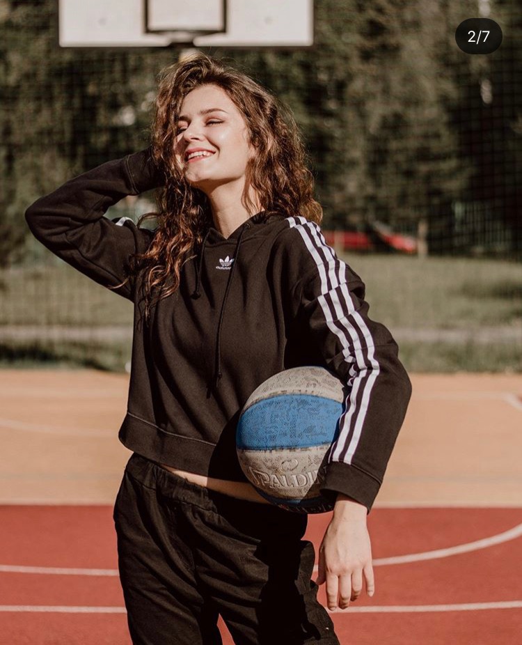 Uśmiechnięta młoda kobieta z kręconymi włosami w czarnej bluzie z kapturem i dresach trzyma zużytą piłkę do koszykówki na tle boiska.