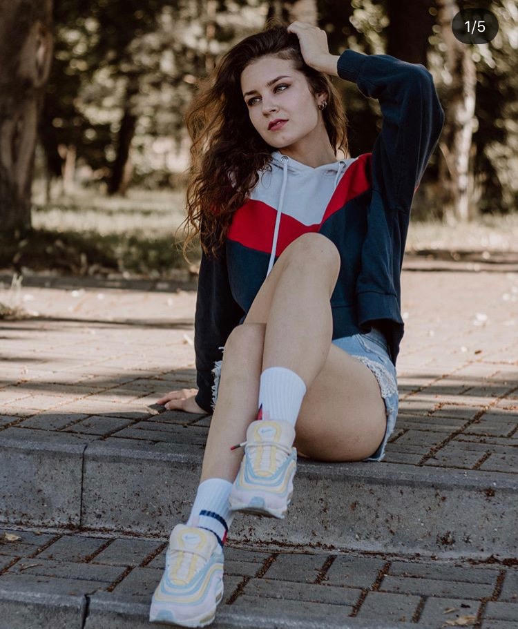 Portret młodej kobiety siedzącej na betonowych schodach w parku, w bluzie z kolorowym wzorem chevron i dżinsowych szortach, z ręką we włosach i pastelowych sneakersach.