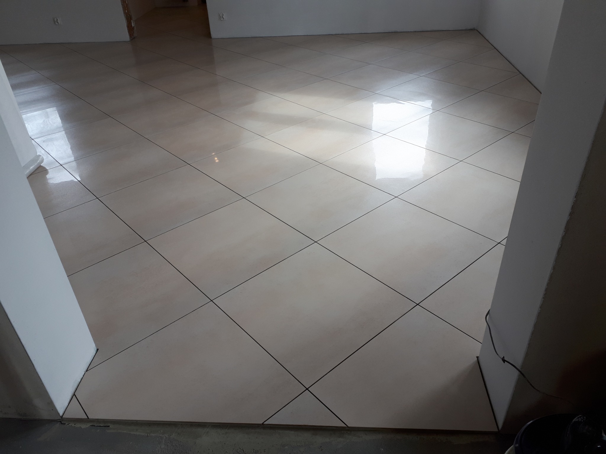 Nowo położona, błyszcząca, beżowa podłoga z płytek ceramicznych ułożonych na skos w pustym pomieszczeniu w trakcie remontu, widoczne fugi i fragment ściany.