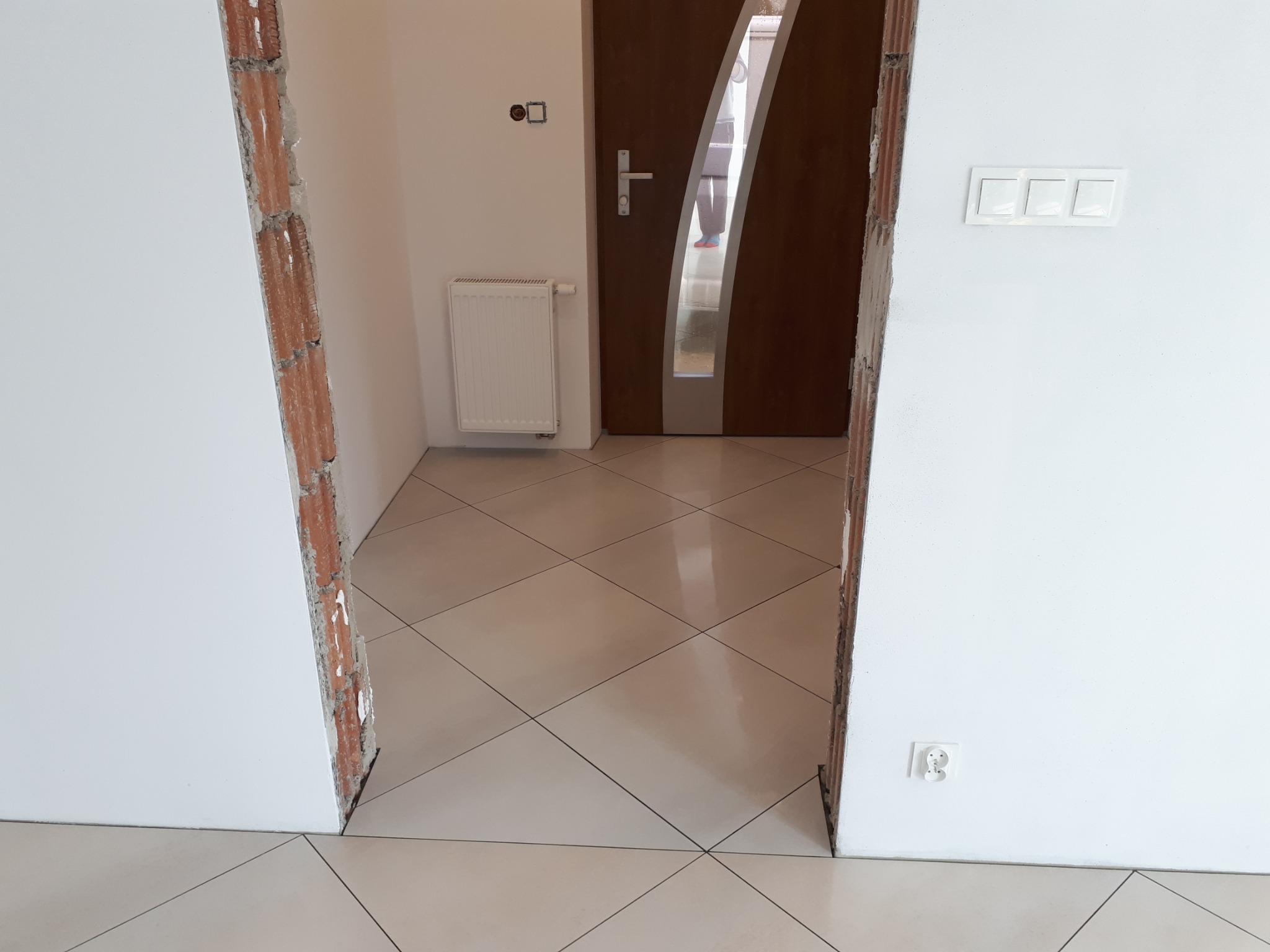 Korytarz w trakcie układania płytek podłogowych, widoczne fragmenty ściany z odsłoniętą cegłą przy wejściu, oraz nowe płytki podłogowe w odcieniach beżu.