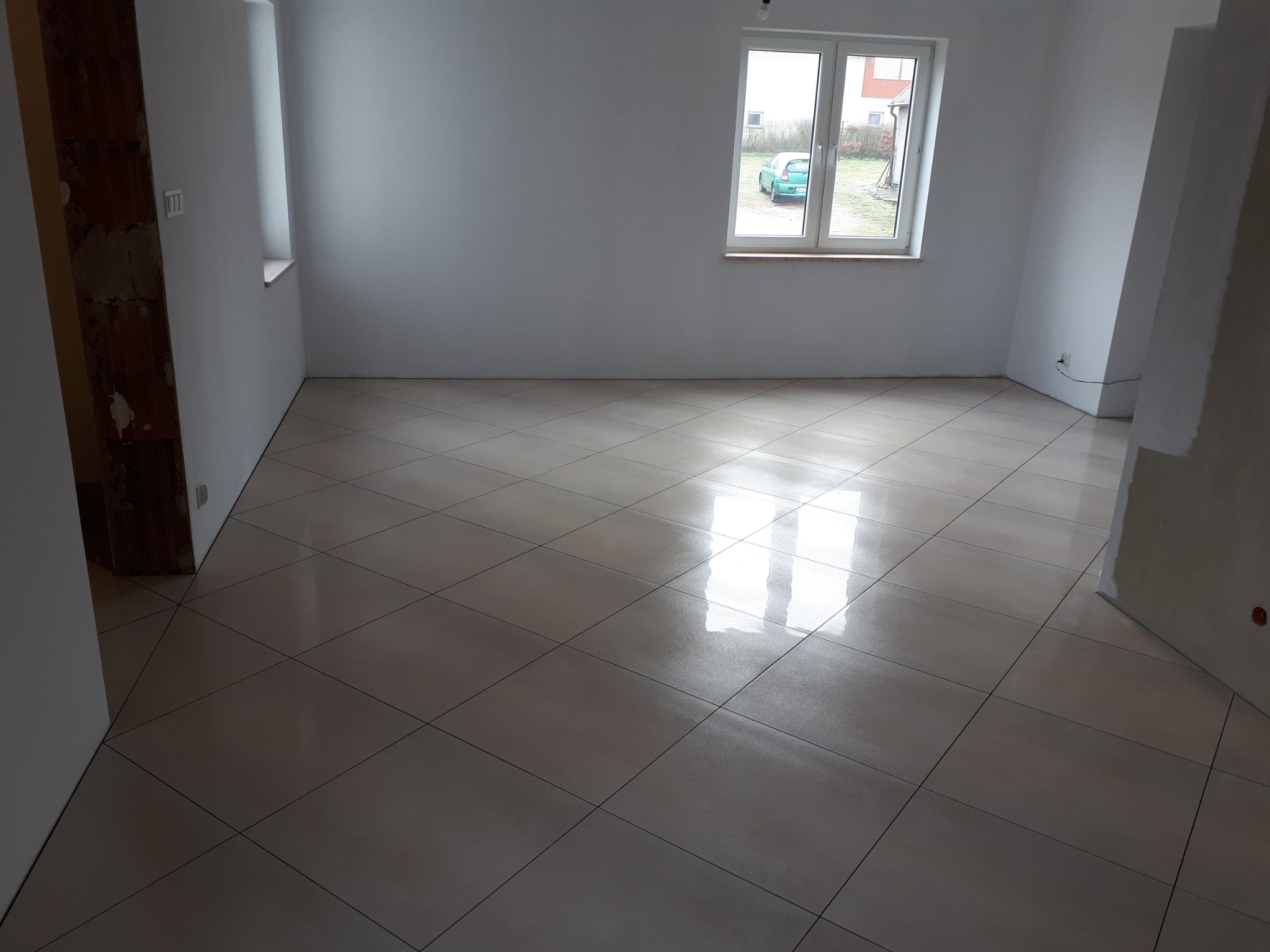 Ułożone płytki podłogowe w nowej łazience, widoczne niedokończone ściany i okno z widokiem na podwórze.