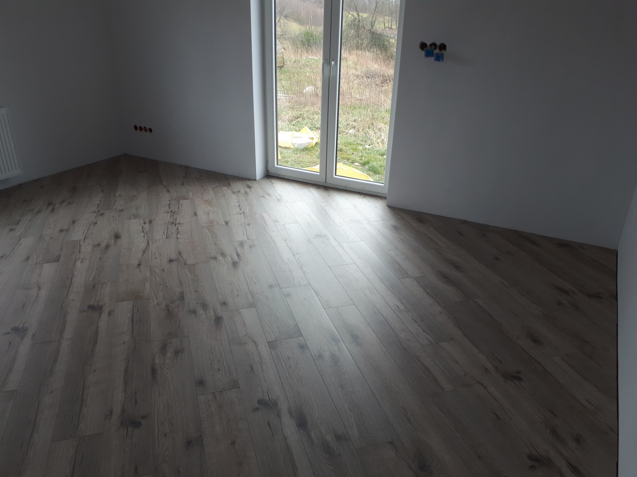 Ułożona podłoga laminowana z wzorem drewna w jasnym odcieniu szarości w pustym pokoju z białymi ścianami, widoczne gniazdka elektryczne i okno balkonowe z widokiem na zewnątrz.