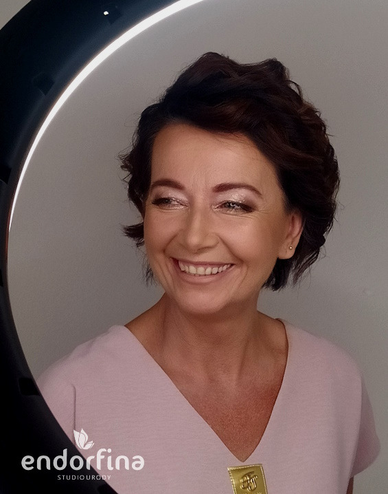 Uśmiechnięta brunetka w różowej bluzce, z delikatnym makijażem oczu i ust, oświetlona lampą pierścieniową, na tle jasnego studia urody.