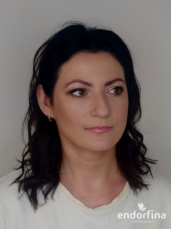 Portret kobiety z delikatnym makijażem, brązowe oczy, błyszczące usta, złote kolczyki, ciemne włosy, logo studia urody w prawym dolnym rogu.