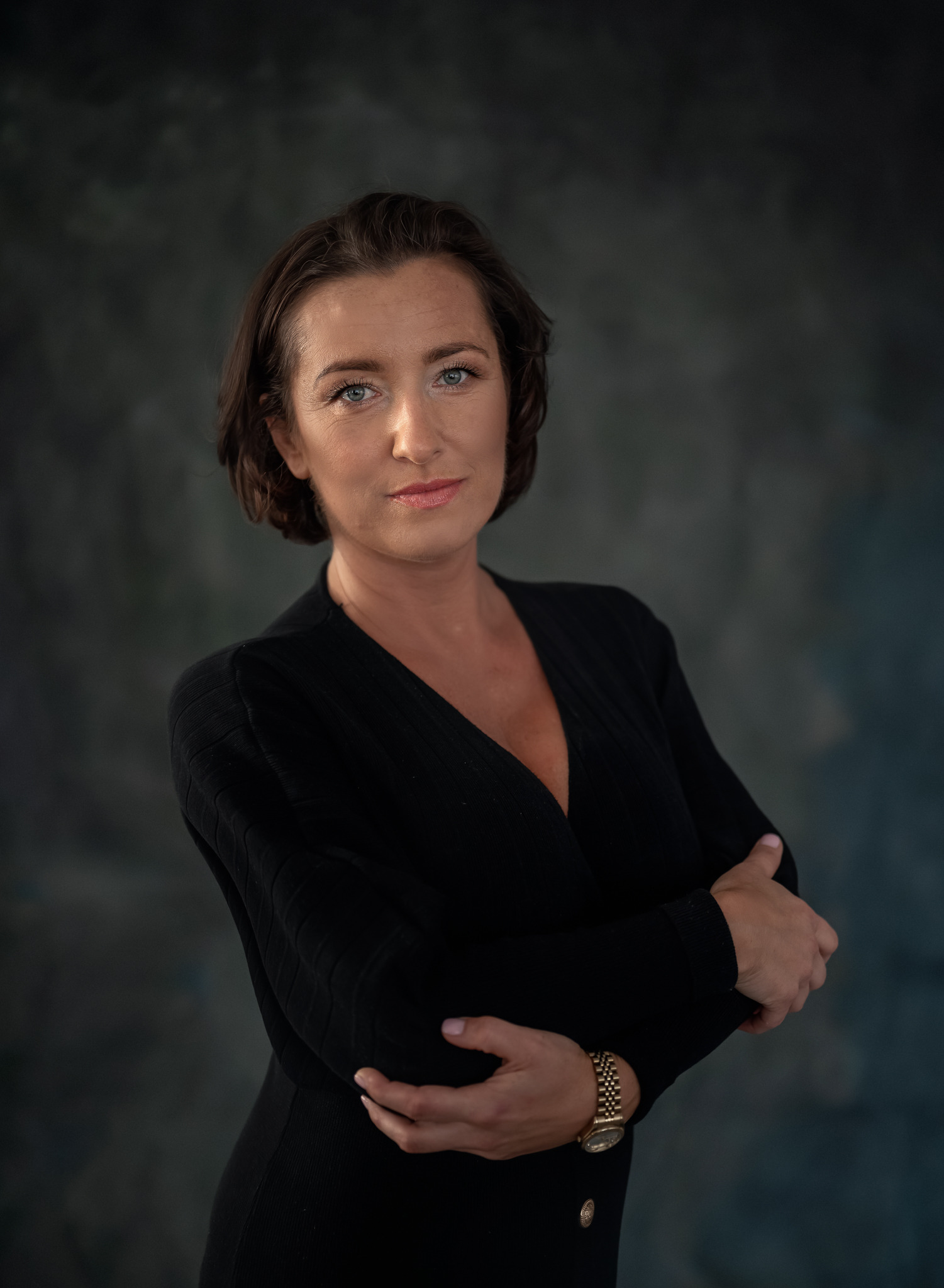 Portret kobiety o niebieskich oczach, w czarnej sukience z rękawami skrzyżowanymi na piersiach, z zegarkiem na ręku, na tle ciemnego, niebieskiego tła.