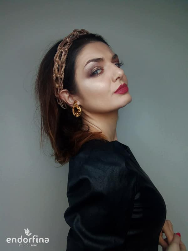 Portret kobiety z wieczorowym makijażem, złotą opaską i kolczykami na tle szarej ściany, z logo studia urody w lewym dolnym rogu.