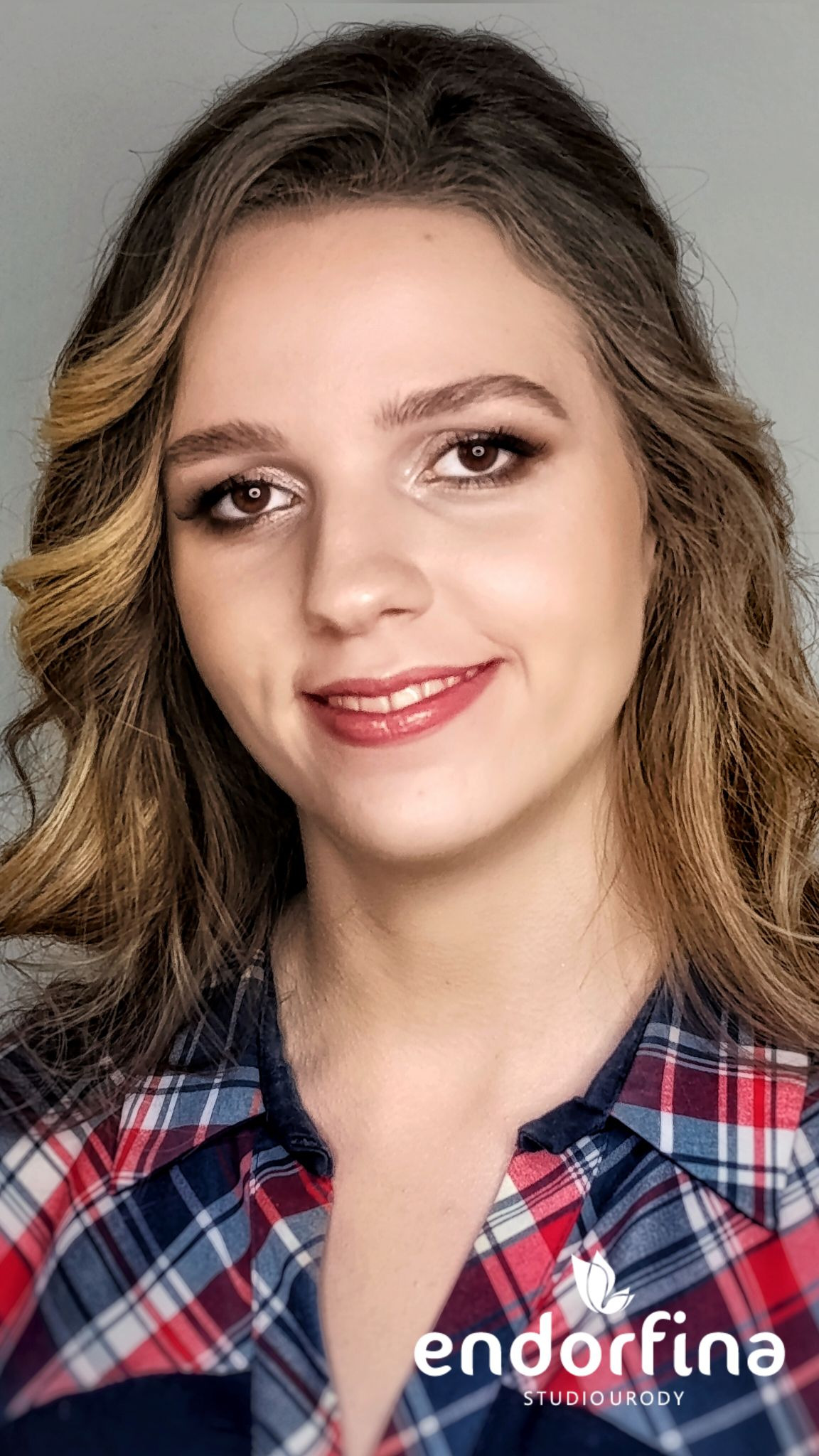 Uśmiechnięta kobieta z delikatnym makijażem oczu i błyszczącymi ustami, w koszuli w kratę. Widoczne logo studia urody w prawym dolnym rogu.