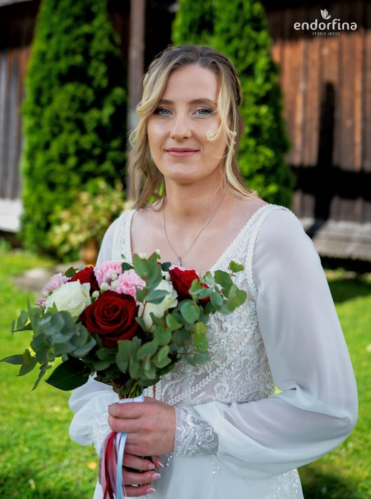 Panna młoda z delikatnym makijażem ślubnym, trzymająca bukiet czerwonych i białych róż, z zielonym tłem. Widoczne logo studia na górze zdjęcia.