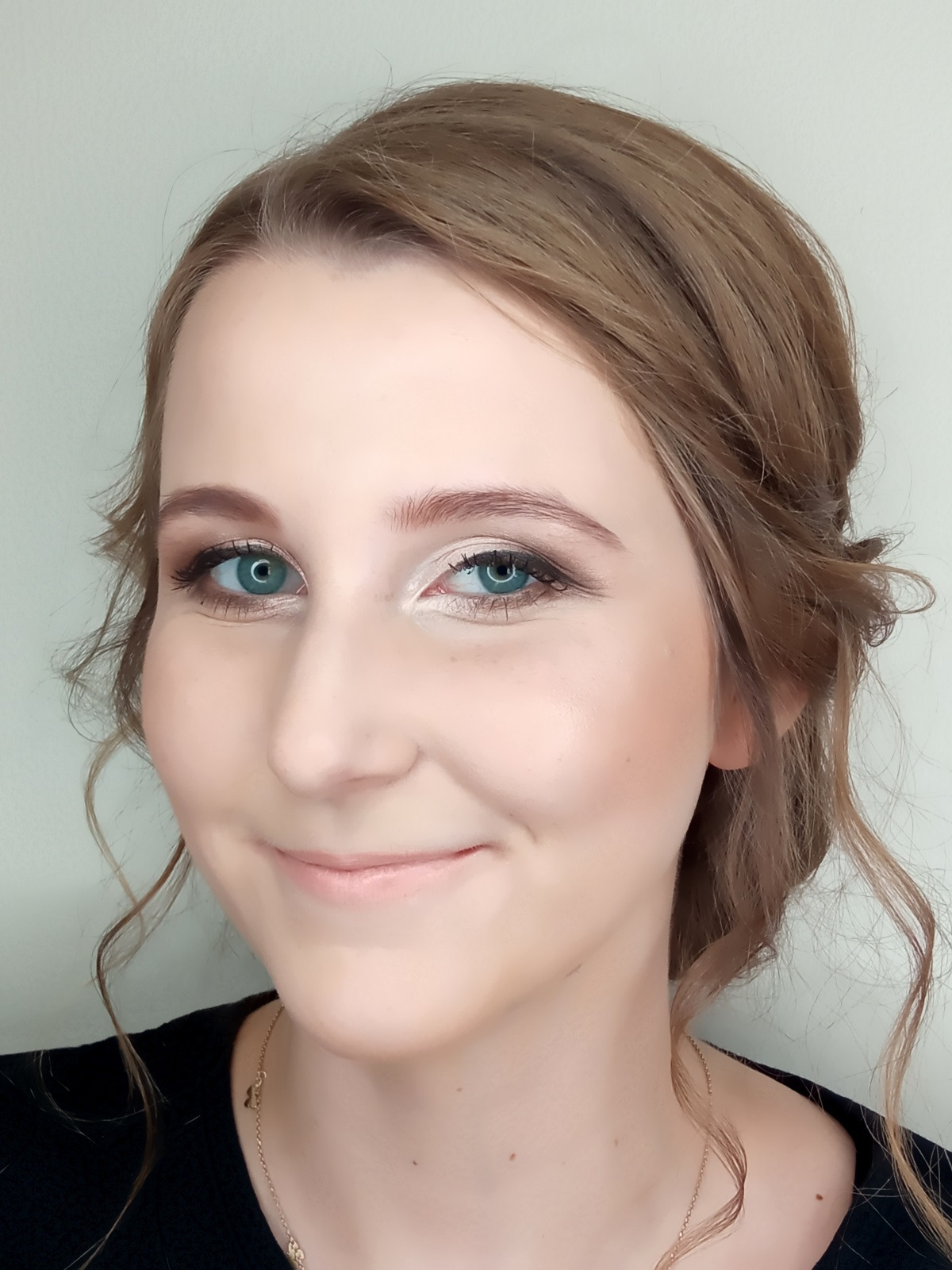 Portret uśmiechniętej kobiety z delikatnym makijażem oczu i ust, upiętymi włosami w odcieniach blondu. Widoczne refleksy światła na kościach policzkowych.