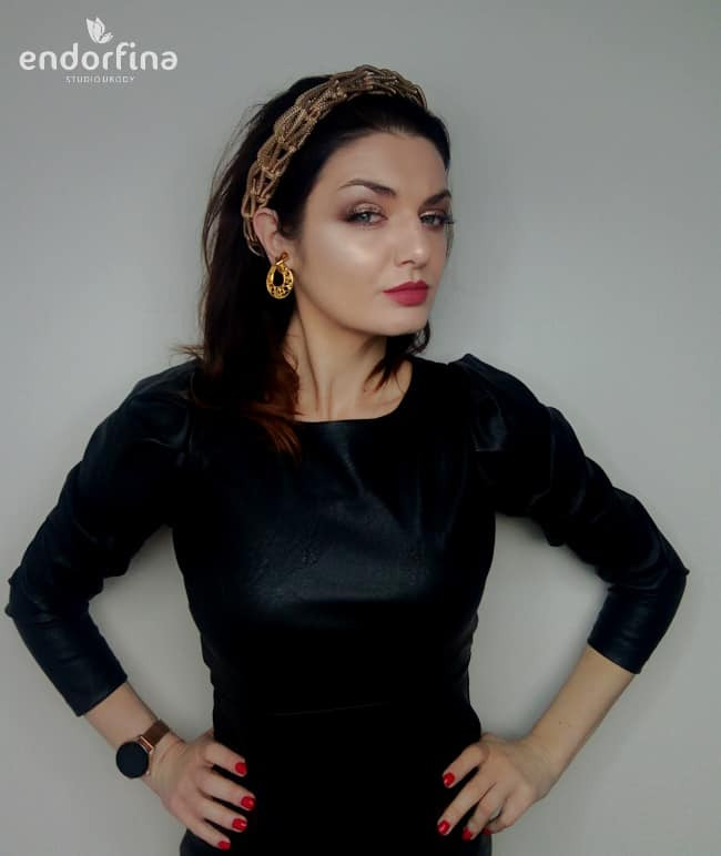 Kobieta w czarnej sukience i złotej opasce, z wyrazistym makijażem oczu i ust, pozuje na jasnym tle. Widoczny zegarek i kolczyki, całość w stylu glamour.