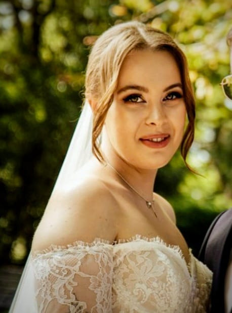 Panna młoda w koronkowej sukni z welonem, subtelny makijaż ślubny podkreśla naturalną urodę, delikatna biżuteria, rozmyte tło zieleni.