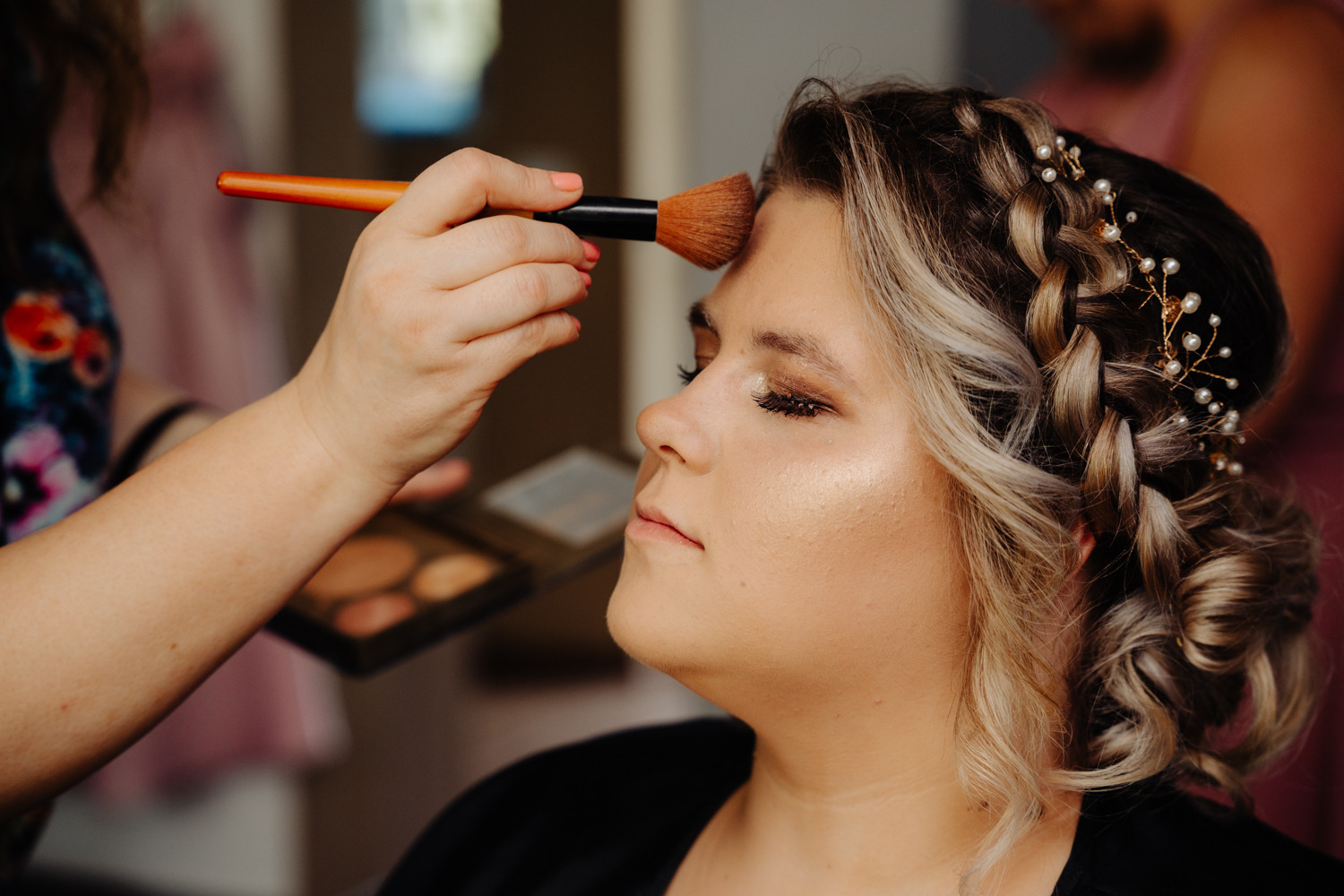 Aplikacja pudru pędzlem na twarz kobiety z upiętymi włosami i ozdobami. Widoczna paleta kolorów i dłoń w trakcie wykonywania usługi. Delikatny makijaż.