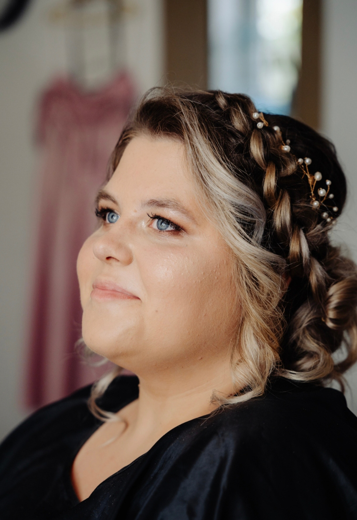 Portret kobiety z upiętymi włosami w warkocze, ozdobionymi perłami. Delikatny makijaż podkreśla niebieskie oczy. W tle widoczna suknia.