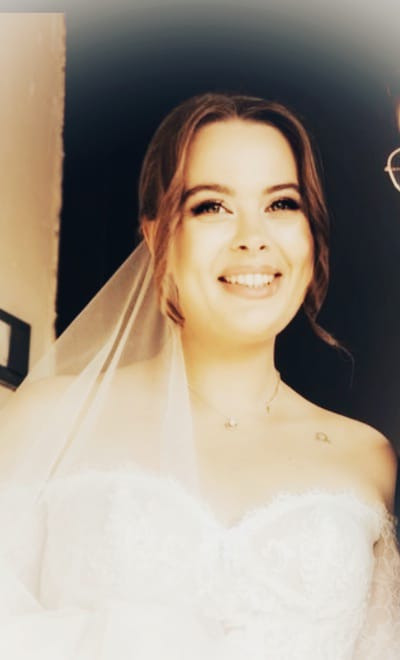 Uśmiechnięta panna młoda w białej sukni z welonem, z delikatnym makijażem i upiętymi włosami, pozuje do zdjęcia w ciemnym pomieszczeniu.