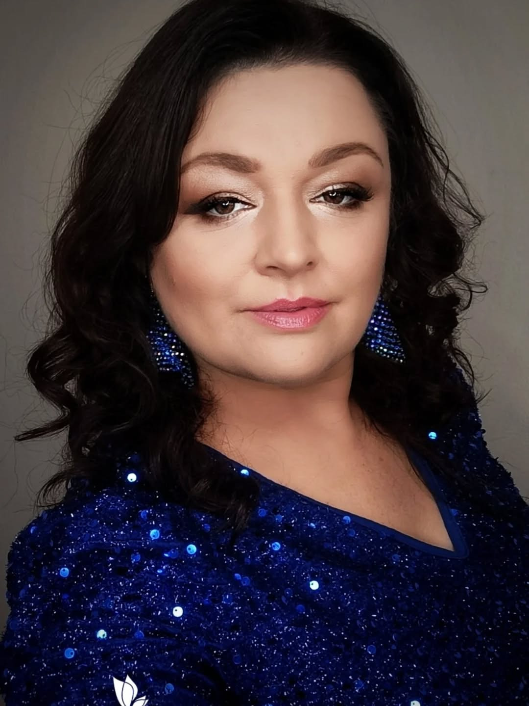 Portret kobiety z ciemnymi włosami w niebieskiej, cekinowej sukience i dopasowanych kolczykach, prezentujący makijaż z brązowymi cieniami i różowymi ustami.