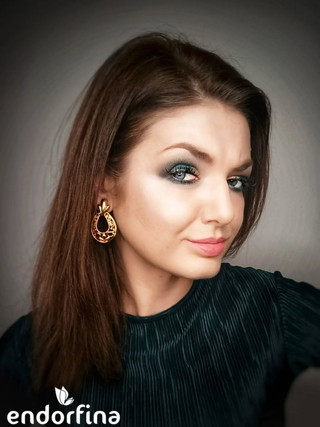 Portret kobiety z profesjonalnym makijażem oczu w odcieniach szarości i zieleni, z widoczną złotą kolczykiem i ciemnozieloną bluzką na szarym tle.