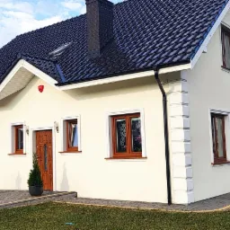 Nowoczesny, parterowy dom z ciemną dachówką, brązowymi oknami i drzwiami, wykończony białą elewacją z dekoracyjnymi elementami, z garażem z boku i zadbanym trawnikiem.