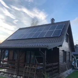 Brązowy dom z drewnianą werandą i panelami słonecznymi na dachu, widok z poziomu trawnika przy zachodzącym słońcu.